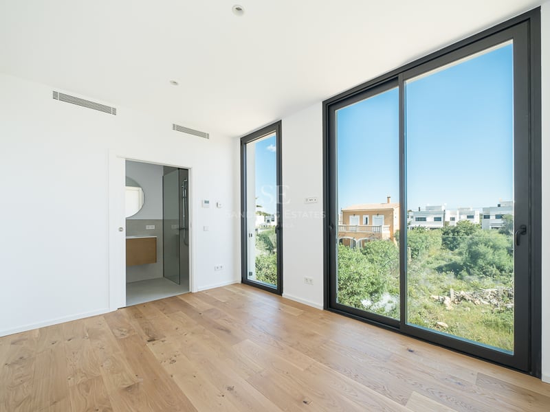Chambre moderne vide avec parquet clair, grandes baies vitrées noires donnant sur le jardin et salle de bain attenante.