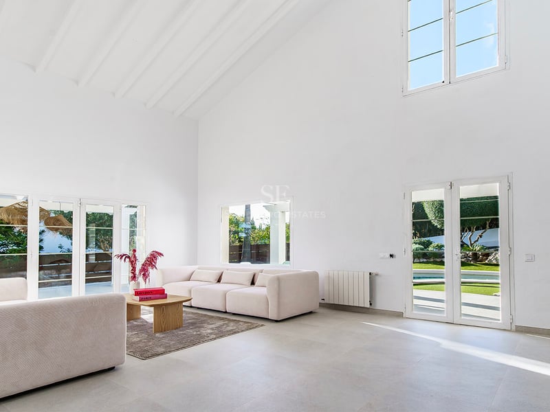 Salon spacieux aux plafonds blancs, canapés beige et portes-fenêtres ouvrant sur le jardin ensoleillé.
