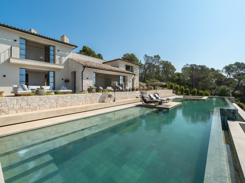 Vue d'une villa de luxe avec une grande piscine à débordement, murs en pierre et bains de soleil face à la forêt.