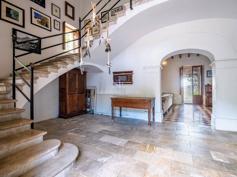 Un grand hall d'entrée avec un escalier en pierre, des meubles anciens en bois et une galerie d'art sous une arche.