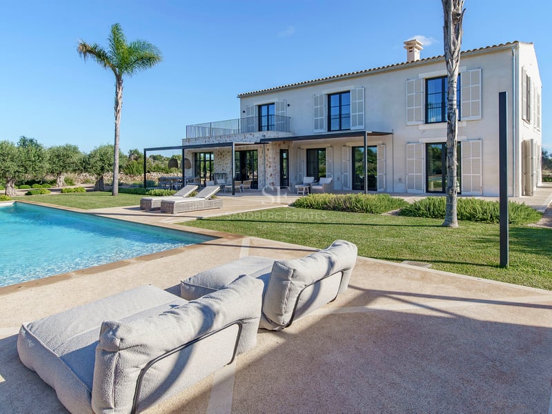 Grande piscine turquoise devant une villa en pierre avec palmiers et pelouse sous un ciel bleu azur.