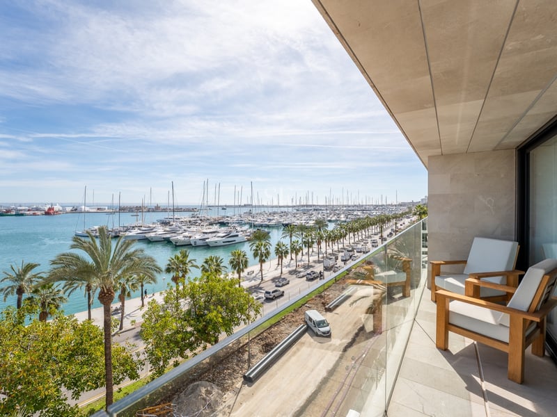 Balcon avec fauteuils en bois et garde-corps en verre surplombant une marina remplie de yachts et de palmiers.