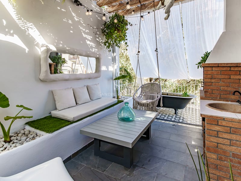 Terrasse blanche avec canapé intégré, balançoire macramé, évier en brique et baignoire noire sous des voiles.