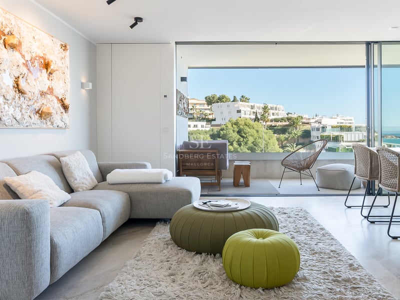 Salón moderno y luminoso con sofá gris y pufs verdes, con ventanales de suelo a techo y vistas al mar.