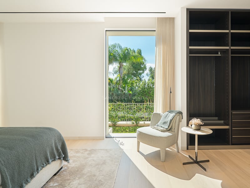 Chambre lumineuse avec un grand dressing en bois sombre, un fauteuil et une vue sur un jardin tropical.