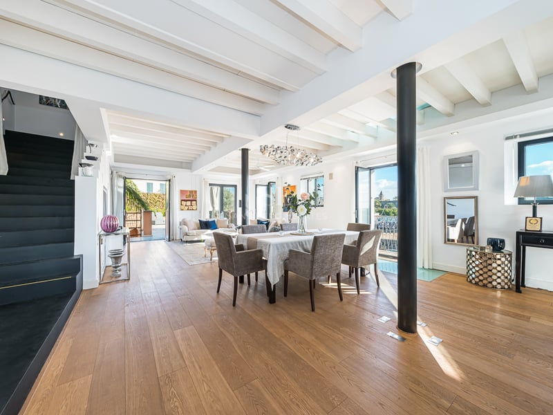 Pièce lumineuse à plan ouvert avec parquet, plafond à poutres blanches, coin repas et escalier noir sur la gauche.