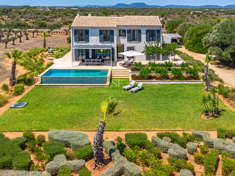 Aerial view of a two-story light gray villa with a terracotta roof, infinity pool, and large green lawn in Mallorca.