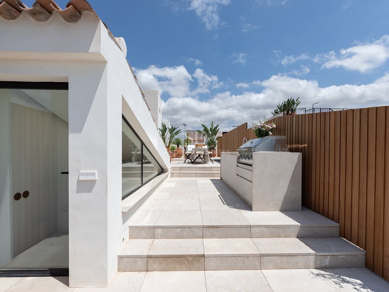 Terraza soleada con cocina de piedra, cerramiento de madera y suelo de piedra caliza bajo un cielo azul.