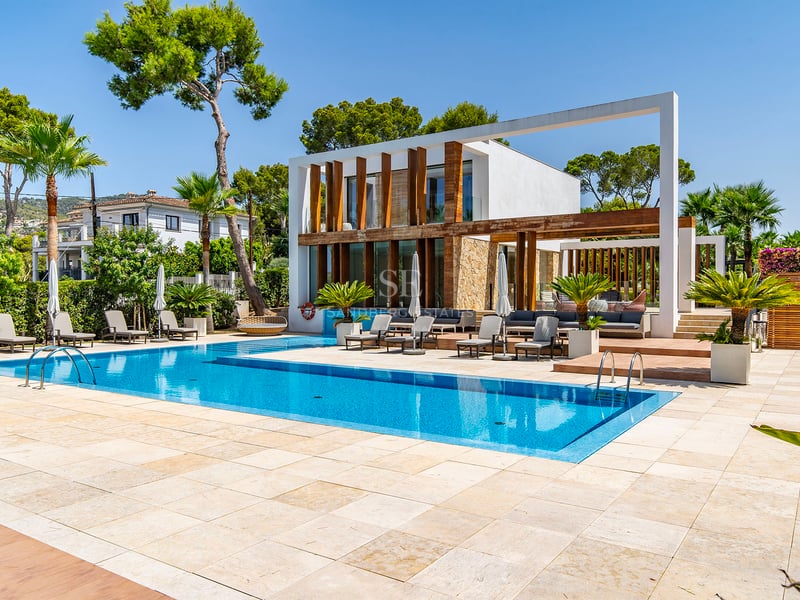 Grande piscine bleue entourée d'une terrasse en pierre claire et d'une villa blanche moderne avec accents en bois.