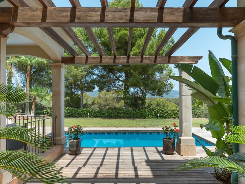Vue d'une pergola en bois sur une piscine bleue entourée d'un jardin luxuriant et d'arbres méditerranéens.