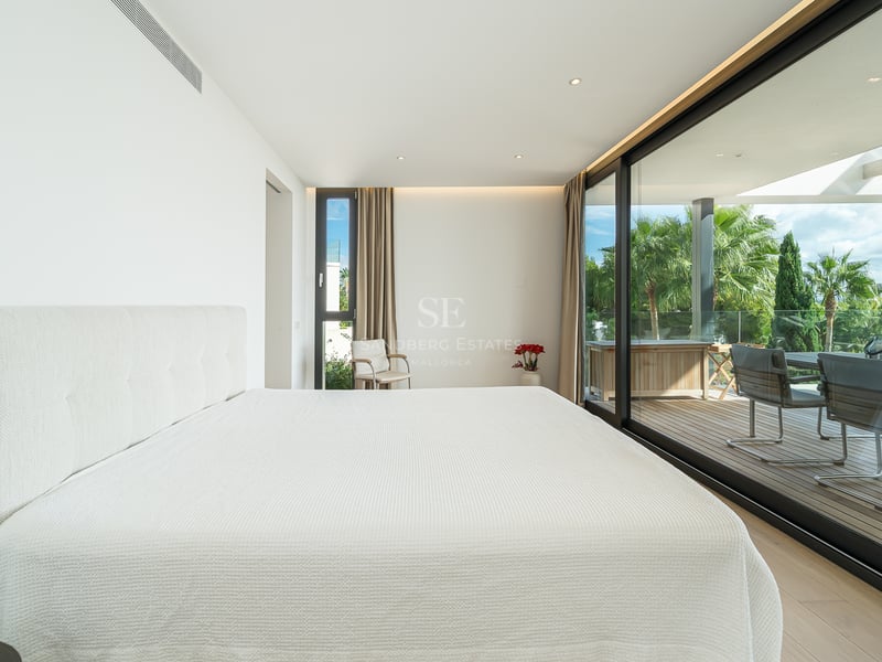Chambre de maître minimaliste blanche avec baies vitrées ouvrant sur une terrasse en bois ensoleillée et des palmiers.
