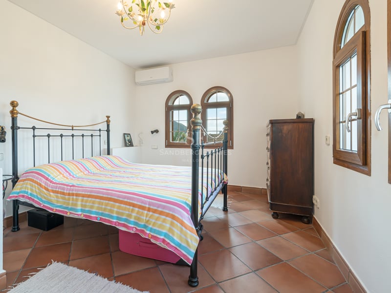 Chambre lumineuse avec sol en terre cuite, lit en fer forgé et fenêtres traditionnelles en bois en forme d'arche.