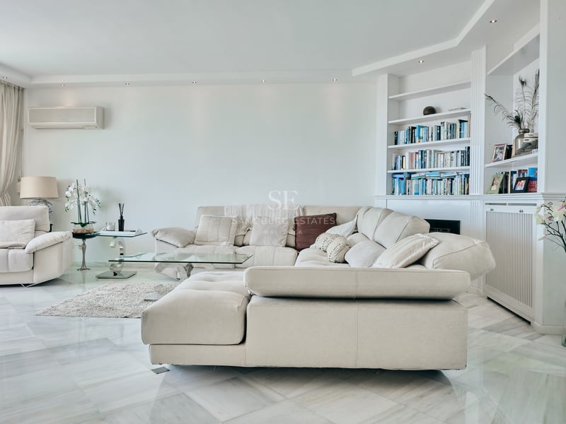 Grand salon avec canapé d'angle beige, sol en marbre poli et bibliothèque intégrée blanche.