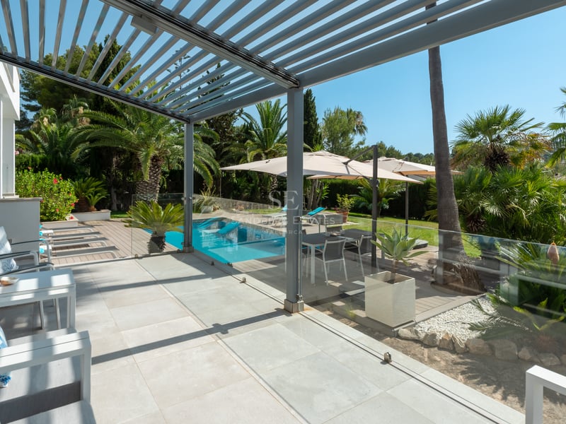Terrasse couverte moderne avec mobilier d'extérieur, vue sur la piscine et un jardin de palmiers sous un ciel bleu.