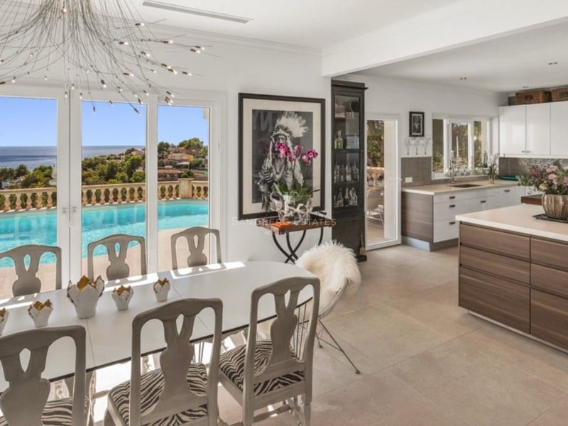 Salle à manger et cuisine modernes avec meubles blancs, donnant sur une piscine à débordement et la mer Méditerranée.