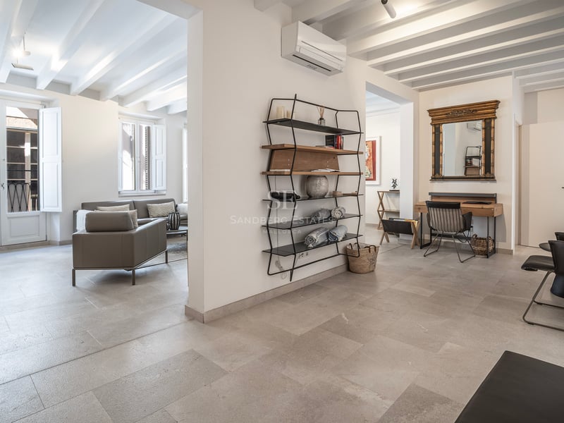 Intérieur moderne avec sol en pierre, poutres blanches, canapé gris et un coin bureau avec miroir doré.