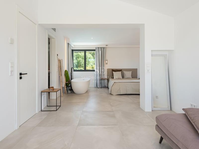 Chambre principale lumineuse avec carrelage grand format, lit minimaliste et baignoire îlot blanche près d'une fenêtre.
