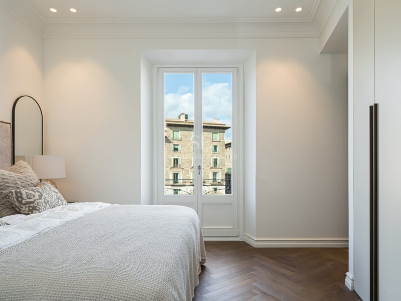 Bright bedroom with dark wood herringbone floors, white built-in wardrobes, and a window overlooking historic buildings.