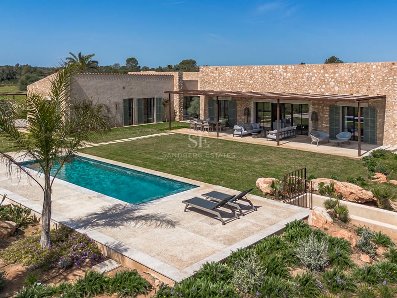 Vista exterior de una villa de piedra con piscina rectangular, césped y terraza bajo pérgola en Mallorca.