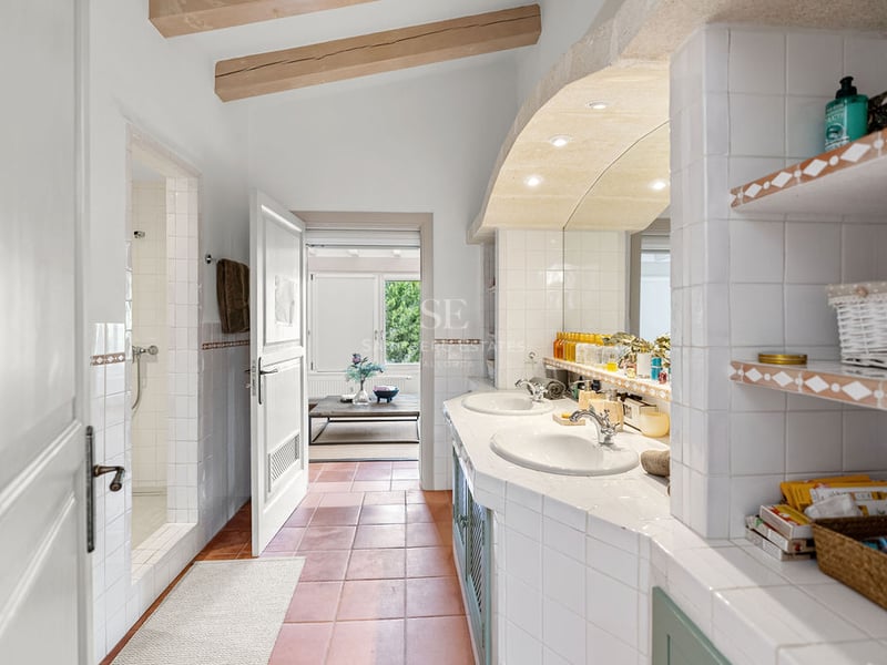Salle de bain spacieuse avec sol en terre cuite, double vasque blanche, poutres en bois et douche à l'italienne.