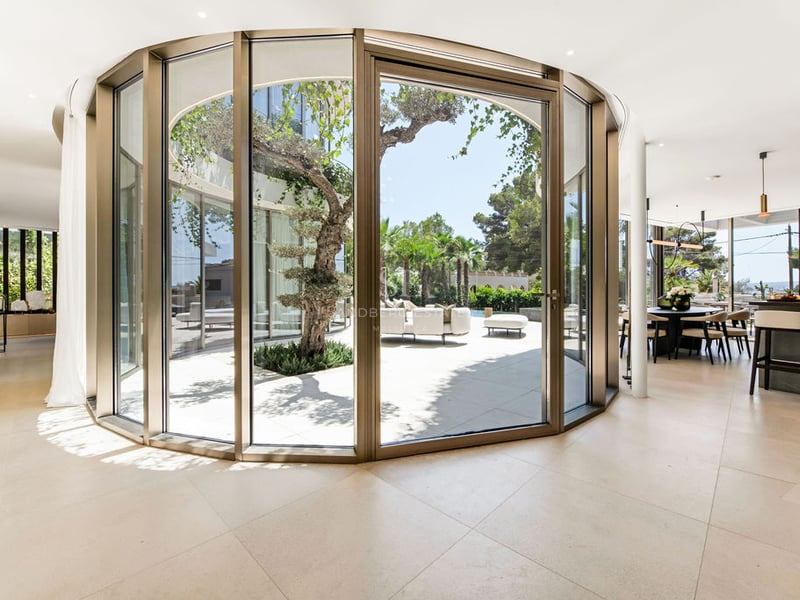 Intérieur d'une villa de luxe avec mur de verre incurvé donnant sur un patio privé avec olivier.