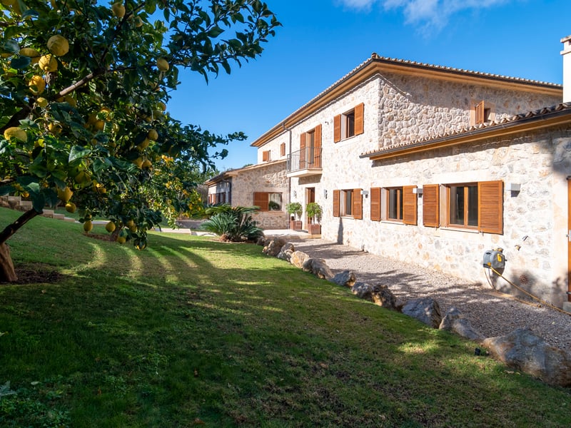Extérieur d'une villa en pierre naturelle avec volets en bois, pelouse et citronniers sous un ciel bleu.