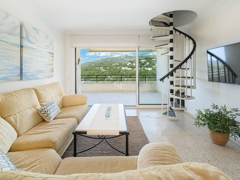 Salon lumineux avec canapé crème, escalier en colimaçon noir et blanc et balcon avec vue sur la montagne.