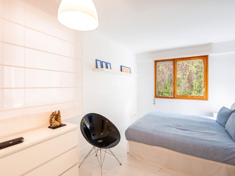 Chambre lumineuse avec grand lit, fenêtre en bois sur verdure, chaise design noire et commode blanche.