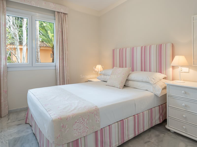 Chambre avec lit double, textiles roses, sol en marbre blanc et fenêtre donnant sur des palmiers.