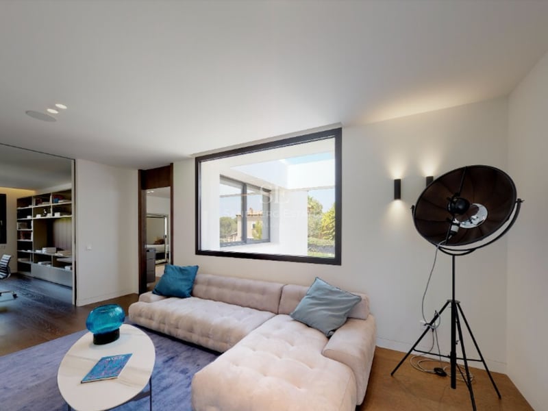 Salon moderne avec canapé capitonné crème, tapis bleu et grande fenêtre donnant sur un patio ensoleillé.
