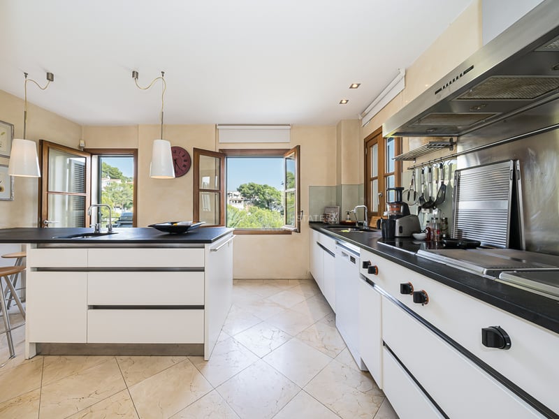 Cuisine contemporaine blanche avec plans de travail sombres, sol en carrelage et fenêtres donnant sur la verdure.