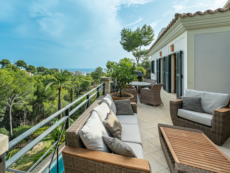 Grande terrasse avec mobilier de salon en rotin et sol en pierre surplombant une vallée verdoyante et la mer.
