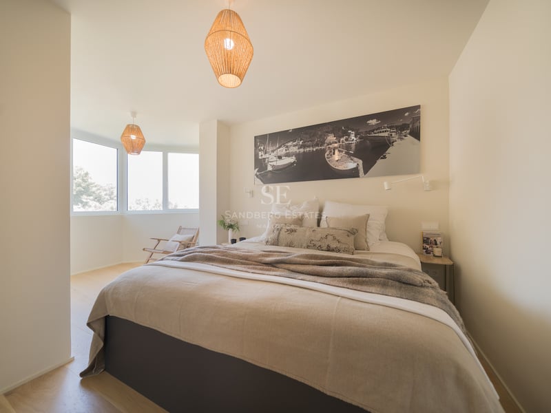 Chambre lumineuse avec un grand lit, des suspensions en osier et une grande fenêtre latérale.