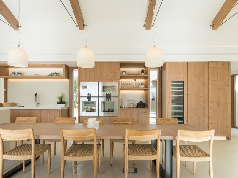 Light wood dining table with woven chairs in front of a modern oak kitchen featuring integrated appliances and wine cooler.