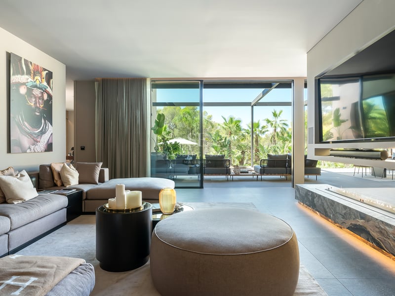 Salon moderne avec canapé d'angle gris, cheminée linéaire et baies vitrées ouvrant sur un jardin tropical.