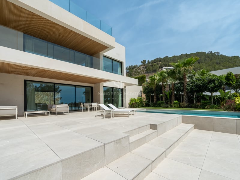 Villa moderne blanche avec grande terrasse en pierre, piscine, garde-corps en verre et collines verdoyantes.