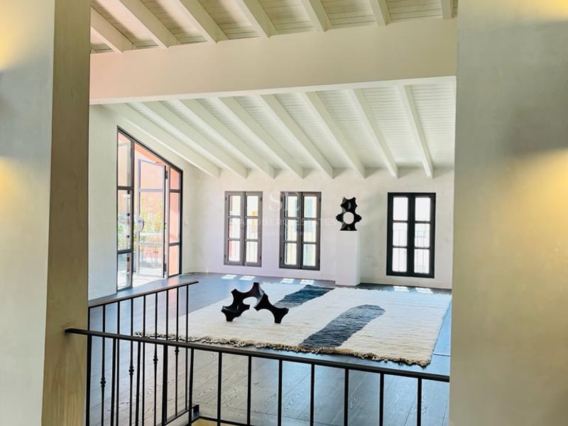 Vue intérieure depuis une mezzanine montrant un salon avec parquet sombre, poutres blanches et fenêtres industrielles.