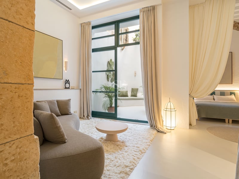 Salon lumineux avec canapé beige incurvé, tapis blanc moelleux et grandes portes vitrées ouvrant sur un patio intérieur.