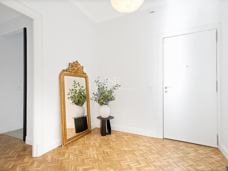 Entrée lumineuse avec murs blancs, parquet en chevrons, un grand miroir doré et une porte blanche.