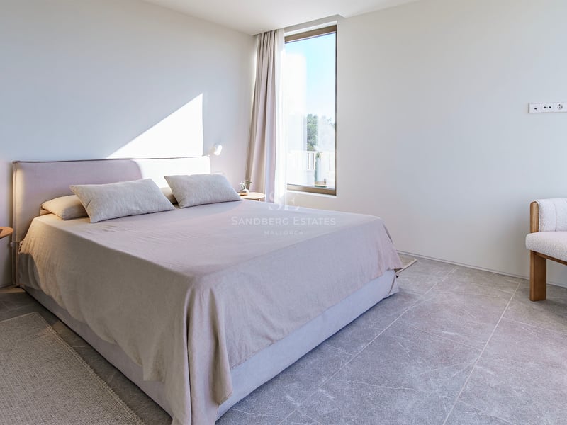 Chambre lumineuse avec carrelage en pierre grise, lit rembourré beige, table de chevet en bois et fauteuil design.