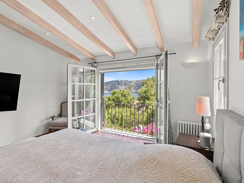 Chambre lumineuse avec poutres en bois et balcon ouvert donnant sur la mer et les collines.