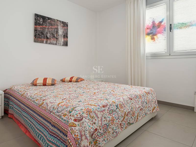 Chambre moderne aux murs blancs, sol en carrelage gris, lit double avec couvre-lit coloré et rideaux blancs.
