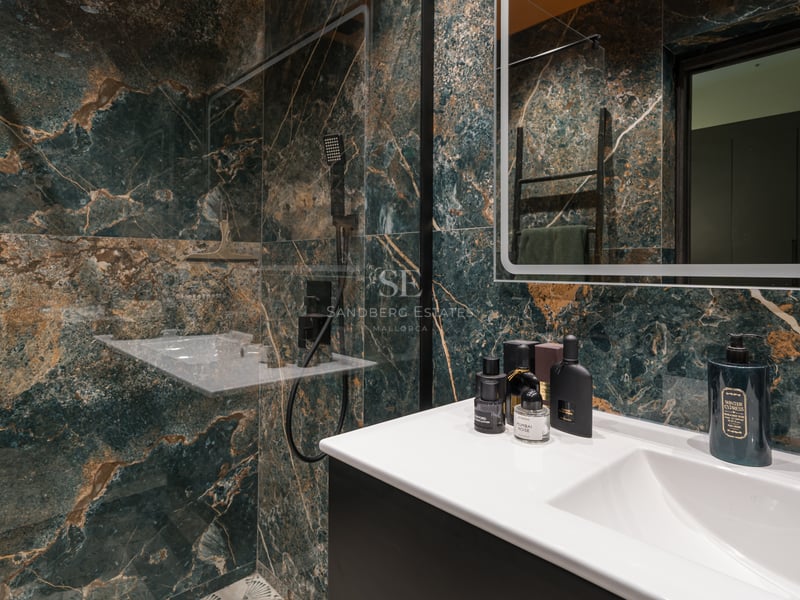 Salle de bains haut de gamme avec murs en marbre vert foncé, veines dorées, vasque blanche et robinetterie noire mate.