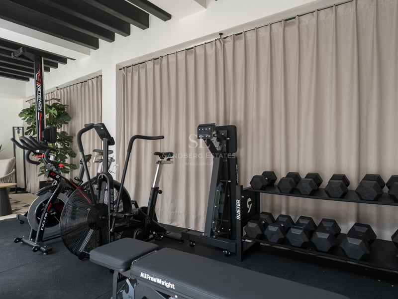Gimnasio moderno con bicicletas estáticas, mancuernas y banco de pesas bajo vigas negras y cortinas beige.