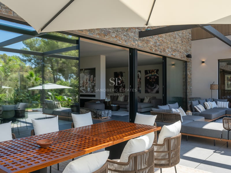 Terrasse moderne avec une grande table en bois, des canapés gris et des baies vitrées ouvrant sur le salon.