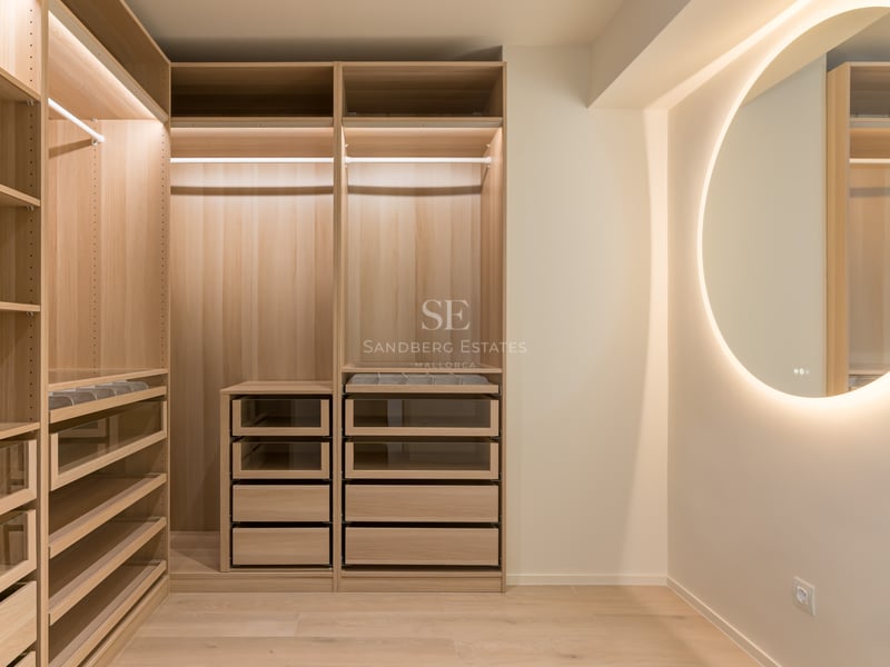 Modern walk-in dressing room featuring light oak cabinetry, glass-fronted drawers, and a large backlit circular mirror.