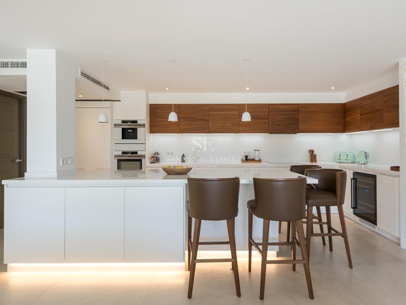 Cuisine moderne avec îlot blanc, tabourets en cuir, placards en noyer et mur en pierre naturelle.