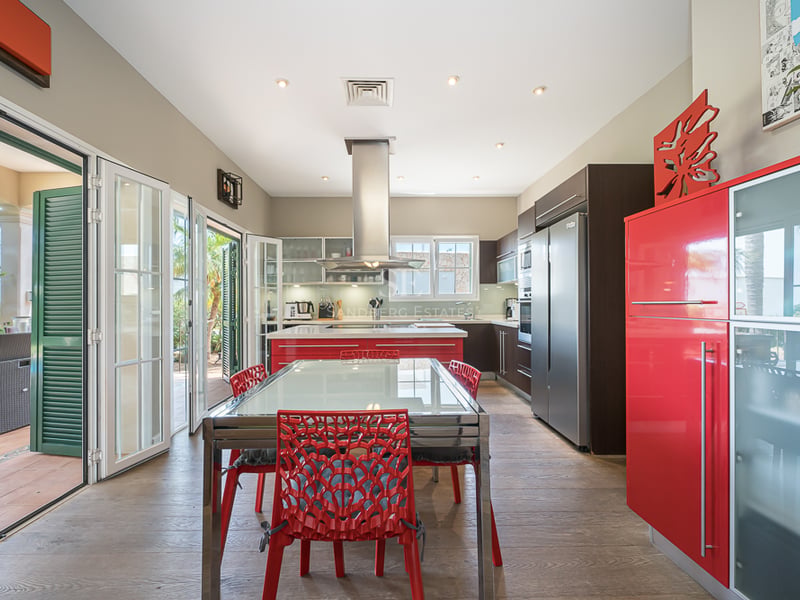 Cuisine lumineuse avec îlot rouge, électroménager en inox et table à manger près des portes vitrées.