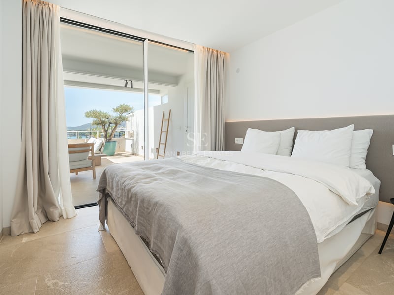 Chambre lumineuse avec grand lit, sol en pierre et baies vitrées ouvrant sur une terrasse ensoleillée.