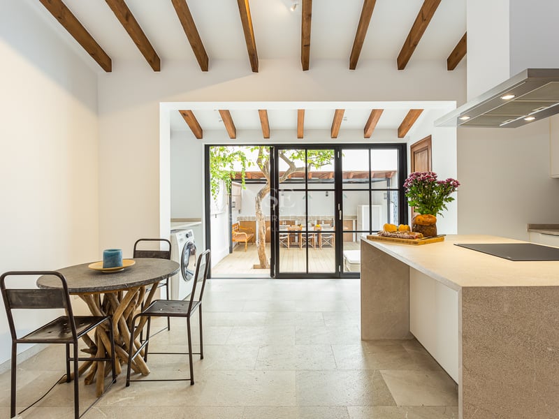 Cuisine lumineuse avec poutres en bois, îlot en pierre et portes vitrées s'ouvrant sur un patio avec un arbre.